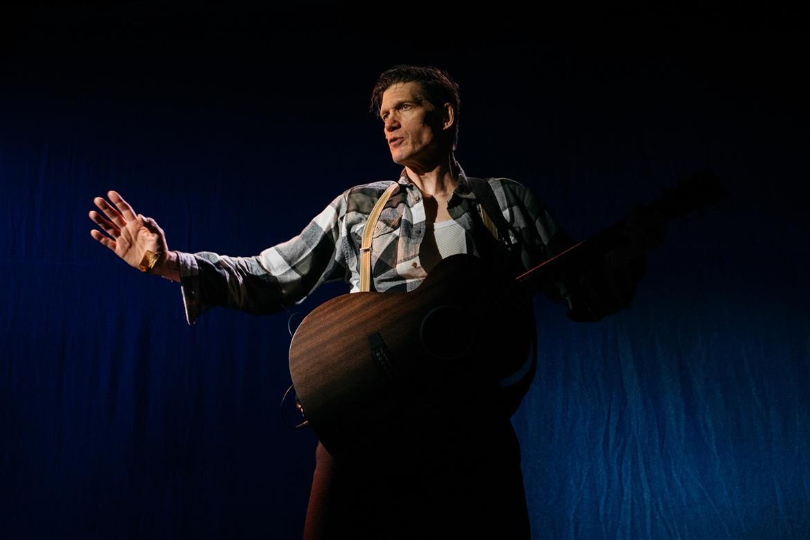 Mies seisoo tummaa taustaa vasten kitara kädessään,