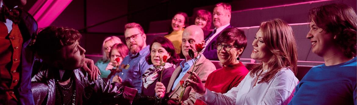 Yleisöä istumassa teatterin katsomossa pinkissä valaistuksessa.